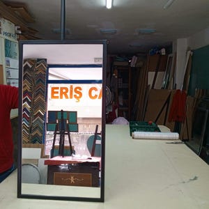 May include: A rectangular mirror with a black frame suspended from the ceiling. The mirror reflects a shop interior with various framed artwork and the text "ERIS CA" visible in the reflection. The shop appears to be a framing business.