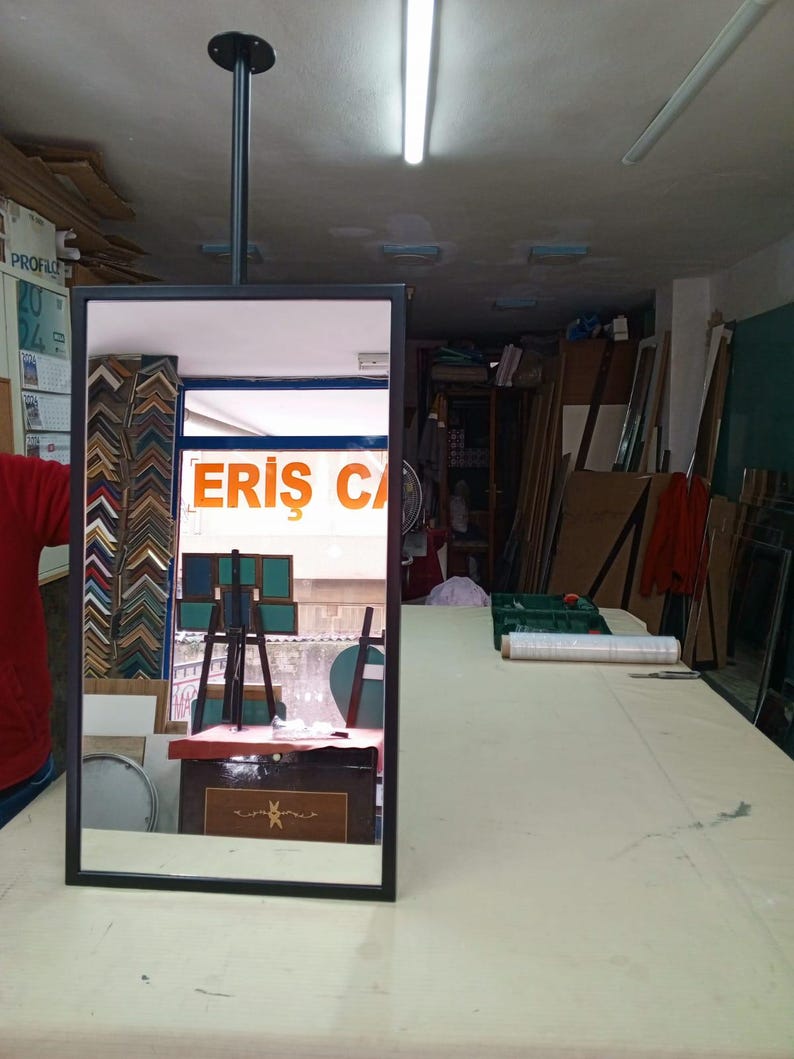May include: A rectangular mirror with a black frame suspended from the ceiling. The mirror reflects a shop interior with various framed artwork and the text "ERIS CA" visible in the reflection. The shop appears to be a framing business.