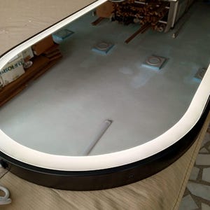 Puede incluir: Un espejo de pared de forma ovalada con un marco negro y una tira de luz LED integrada que recorre el per&iacute;metro interior. La superficie del espejo es reflectante y muestra una vista clara del entorno y de la fuente de luz.
