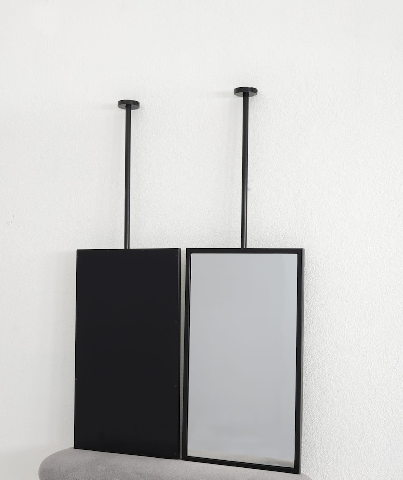 May include: Two rectangular mirrors suspended from the ceiling by black rods. One is a solid black panel, the other a reflective surface. The mirrors are mounted on a grey upholstered bench against a white wall. Each mirror is approximately 30 cm wide.