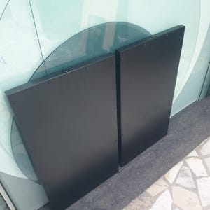 May include: Two black, rectangular panels with rounded edges, possibly for a modern door or wall feature. The panels are positioned upright, with visible rivets along the edges. The background includes glass panels and a light-coloured floor.