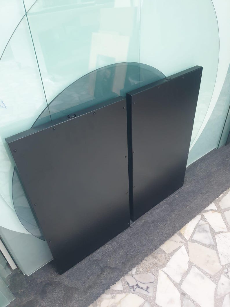 May include: Two black, rectangular panels with rounded edges, possibly for a modern door or wall feature. The panels are positioned upright, with visible rivets along the edges. The background includes glass panels and a light-coloured floor.