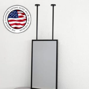 May include: A rectangular mirror with a black frame, suspended from the ceiling by two black rods. The mirror is positioned against a white wall. The image includes a circular graphic with the American flag and text.