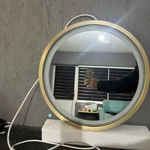 Puede incluir: Un espejo de pared circular con un marco de metal en tono dorado cepillado y una tira de luz LED integrada a lo largo del borde interior. El espejo incluye un cable de alimentaci&oacute;n blanco que sale por la parte superior y descansa sobre una base de espuma blanca.