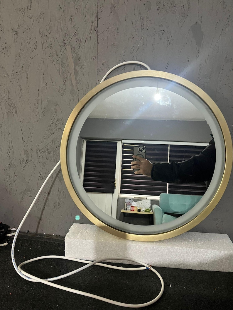 Puede incluir: Un espejo de pared circular con un marco de metal en tono dorado cepillado y una tira de luz LED integrada a lo largo del borde interior. El espejo incluye un cable de alimentaci&oacute;n blanco que sale por la parte superior y descansa sobre una base de espuma blanca.