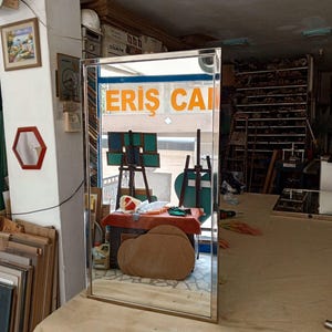 May include: A rectangular mirror with a thin, gold-coloured frame. The mirror reflects a workshop interior, including a table, easels, and the text "ERIS CAI" in orange. The mirror is positioned on a light-coloured surface.