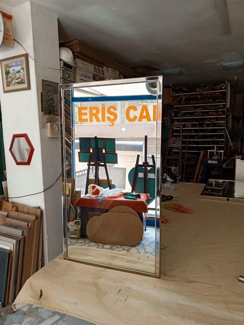 May include: A rectangular mirror with a thin, gold-coloured frame. The mirror reflects a workshop interior, including a table, easels, and the text "ERIS CAI" in orange. The mirror is positioned on a light-coloured surface.