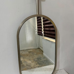 Puede incluir: Un espejo de forma ovalada con un marco de metal plateado. El espejo est&aacute; unido a una varilla cil&iacute;ndrica vertical que se conecta a una placa de montaje circular en la parte superior, lo que permite colgar el espejo desde el techo.