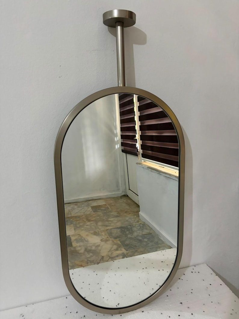 Puede incluir: Un espejo de forma ovalada con un marco de metal plateado. El espejo est&aacute; unido a una varilla cil&iacute;ndrica vertical que se conecta a una placa de montaje circular en la parte superior, lo que permite colgar el espejo desde el techo.