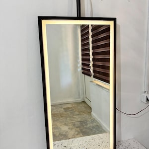 Puede incluir: Un espejo rectangular montado en la pared con un marco negro y una tira de luz LED integrada a lo largo del per&iacute;metro interior. El espejo cuenta con un peque&ntilde;o bot&oacute;n de encendido t&aacute;ctil circular en la parte inferior central y est&aacute; suspendido por una varilla de metal negro.