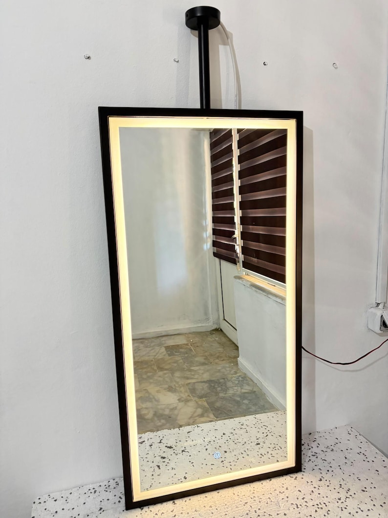 Puede incluir: Un espejo rectangular montado en la pared con un marco negro y una tira de luz LED integrada a lo largo del per&iacute;metro interior. El espejo cuenta con un peque&ntilde;o bot&oacute;n de encendido t&aacute;ctil circular en la parte inferior central y est&aacute; suspendido por una varilla de metal negro.