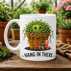 May include: A white ceramic mug featuring a graphic of a green string of pearls plant in a terracotta pot with a face, a small pink worm holding a tiny watering can, and the black text HANG IN THERE printed at the bottom of the design.