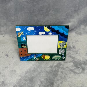 May include: A rectangular picture frame featuring a painted scene of a boat on water with various animals including a giraffe, lion, elephant, and zebra. The frame has a dark green border, a blue sky with a yellow moon, and white clouds.