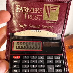 Könnte beinhalten: Eine weinrote Kunstleder-Tasche für einen Taschenrechner mit dem Farmers Trust-Logo in Gold. Der Rechner zeigt "00080080" auf schwarzem Hintergrund an. Die Hülle enthält auch eine Kreditkarte.