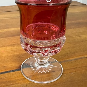 Puede incluir: Una copa de cristal rojo rubí con base y tallo transparentes y estampados. La parte superior del cristal es de color rojo intenso, mientras que la base y el tallo son transparentes. La copa se apoya sobre una base redonda y transparente.