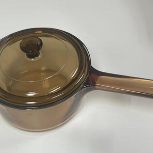 Puede incluir: Cacerola de vidrio color ámbar con tapa a juego y un mango largo. La cacerola es redonda con una tapa de vidrio transparente y un pomo marrón oscuro. El mango también es de color ámbar.