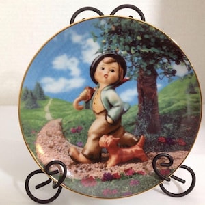 May include: A porcelain plate with a gold rim features a painting of a young boy walking with a dog on a path through a green meadow. The boy is wearing a blue jacket and brown pants. The dog is brown and white. The background is a blue sky with white clouds.