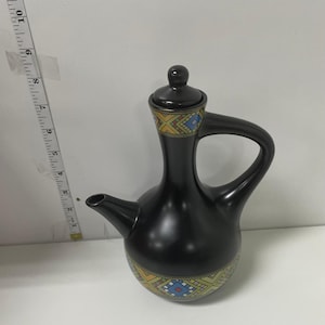 May include: A black ceramic coffee pot with a handle and spout. The pot has a lid and is decorated with a geometric pattern in yellow, blue, and orange. The pot is approximately 23 cm tall.