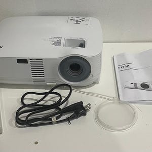 May include: A white NEC VT700 portable projector with a lens, control panel, and ventilation grilles. Included are a gray remote control, a black power cord, a circular translucent lens cap, and a printed manual labeled NEC Portable Projector VT700.