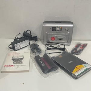 May include: A silver Kodak EasyShare printer dock series 3 with a black power adapter, a user guide, a black ink cartridge, a black paper tray, and two black connection cables. The printer dock features a central circular button and a front slot.