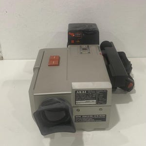 Puede incluir: Cámara de video AKAI VC-X11 vintage en gris y negro. La cámara tiene una lente, un asa y botones. El texto "AKAI VC-X11" es visible en la parte frontal. Un equipo de video clásico.