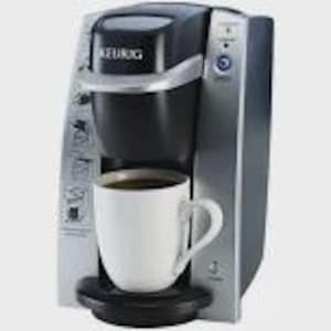 May include: A black and silver Keurig single-serve coffee maker with a white mug filled with coffee. The coffee maker has a digital display and a power button. The mug is placed on a black drip tray.