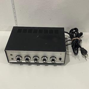 Puede incluir: Amplificador Realistic MPA-20 120V/12V de estado sólido P.A. negro y plateado. El amplificador tiene múltiples perillas para el volumen y el control de tono. El cable de alimentación es negro y está enrollado.