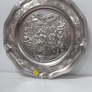May include: A silver metal wall plate with an embossed design of a group of people in a village setting. The plate has a decorative border with a scalloped edge.