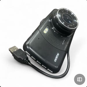 Può includere: Una dash cam nera con obiettivo argentato e cavo USB. La fotocamera ha la scritta "CHORTAU" stampata sul lato. L'obiettivo è circolare e prominente, e c'è una piccola luce sulla parte superiore.