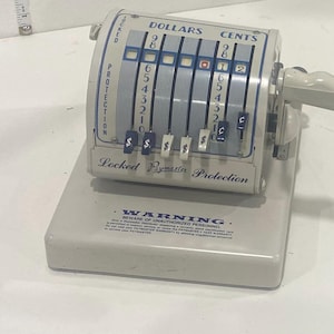 May include: A vintage, off-white cash register with blue and white accents. The register displays dollar and cent amounts, with a handle on the side. The text "Locked Remittance Protection" is visible. A warning label is printed on the base.