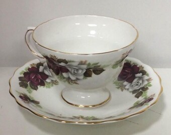May include: White teacup and saucer set with a floral rose design in shades of burgundy, white, and green. The cup and saucer have gold trim. The saucer has a scalloped edge.