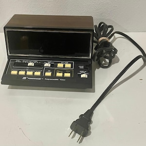 Puede incluir: Un temporizador programable Powerhouse negro y marrón vintage con una pantalla digital. El temporizador tiene múltiples botones y controles deslizantes para configurar la hora y otras funciones. Un cable de alimentación negro con un enchufe se extiende desde el dispositivo.