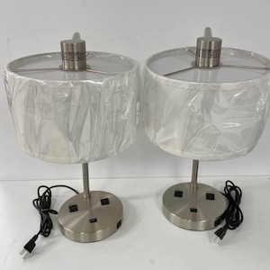 May include: Two matching table lamps with white fabric shades, cylindrical brushed nickel bases, and USB ports. Each lamp has a black power cord and a silver-coloured stem. The shades are covered in protective plastic.