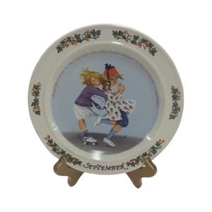 May include: A decorative plate from the Sarah Stilwell Weber Calendar Collection. The plate features a charming illustration of two children, one on roller skates, with the word "September" at the bottom. The plate is displayed on a wooden stand.