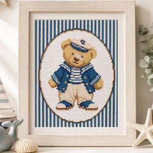 Pode incluir: Um bordado em ponto cruz emoldurado apresentando um urso de peluche castanho claro vestido com um traje de marinheiro azul e branco com um boné a condizer. O urso está centrado numa moldura oval sobre um fundo de riscas verticais azuis e brancas.