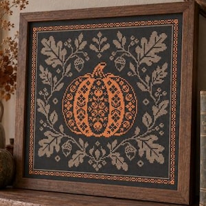 May include: A square framed cross-stitch embroidery featuring a central orange pumpkin with intricate floral patterns, surrounded by a border of gray oak leaves and acorns on a dark background, encased in a dark brown wooden frame.