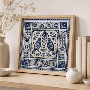 Pode incluir: Uma peça de bordado em ponto cruz quadrada emoldurada em madeira clara, apresentando um padrão geométrico em azul escuro e creme com dois pássaros voltados para um motivo floral central, rodeado por uma borda decorativa de pequenos quadrados.