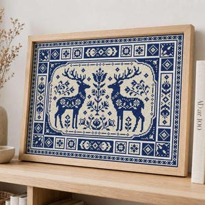 Könnte beinhalten: Eine rechteckige gerahmte Kreuzstichstickerei mit einem symmetrischen Design aus zwei dunkelblauen Hirschen, die einem zentralen Blumenmotiv auf cremefarbenem Grund gegenüberstehen, umrahmt von einer geometrischen Bordüre in Blau und Creme.