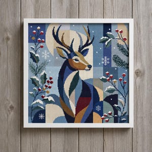 May include: A framed cross-stitch embroidery featuring a stylized deer in shades of blue, brown, and tan. The design includes holly branches with red berries, pine needles, and white snowflakes against a geometric blue and beige background.