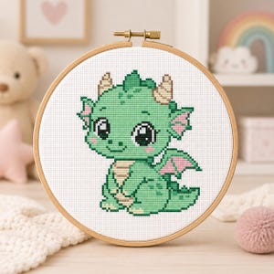 Può includere: Un progetto di ricamo a punto croce raffigurante un drago dei cartoni animati con pelle verde chiaro, ali interne rosa e corna color beige. Il disegno è centrato su tessuto Aida bianco e inserito in un telaio da ricamo in legno circolare.
