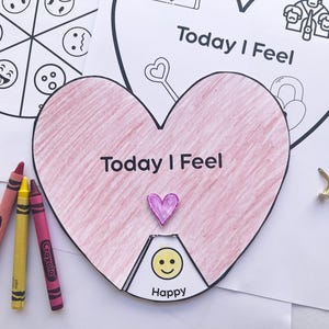 Op de afbeelding: Een hartvormig papieren knutselwerk met de tekst Today I Feel, voorzien van een draaibaar wielsegment met een geel smiley-emoji. Het hart is ingekleurd met roze krijt en bevat een kleine paarse hartvormige uitsparing, drie krijtjes en een metalen splitpen.