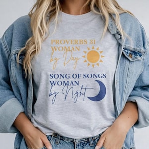 Puede incluir: Camiseta gris claro con el texto "PROVERBIOS 31 WOMAN by Day" y "SONG OF SONGS WOMAN by Night". El diseño incluye un sol amarillo y una luna creciente azul. La persona lleva una chaqueta vaquera.