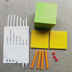 May include: Base ten blocks in various colours for educational use. The set includes a green cube, two yellow squares, orange rods, and small red cubes. A white card with place value labels is also visible.