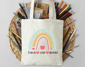 Può includere: Una borsa tote in tela bianca con un design a forma di arcobaleno e le parole "Teach Love Inspire" stampate sul davanti. L'arcobaleno è composto da strisce gialle, arancioni e rosse con una mela rossa al centro. La borsa ha due manici ed è circondata da matite colorate.