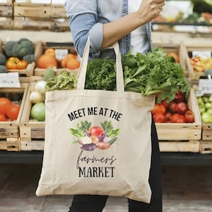May include: A white canvas tote bag with the text "Meet me at the farmers market" and a watercolor illustration of various vegetables and herbs.