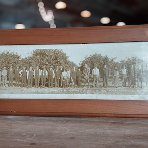 Puede incluir: Una fotografía en blanco y negro enmarcada de un grupo de personas de pie frente a una fila de árboles. Las personas están todas vestidas con atuendos formales y parecen ser de principios del siglo XX.