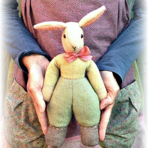 Peut inclure: Un lapin en peluche fait main avec une tenue verte et marron et un nœud papillon rose. Le lapin est tenu par une personne portant une robe à imprimé floral.