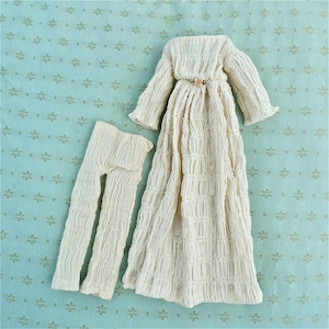 May include: A white, ruffled dress and matching pants for a doll. The dress has a gathered bodice and long sleeves. The pants are loose fitting and have a straight leg.