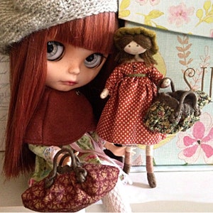 Op de afbeelding: Twee poppen, een met lang rood haar dat een bruine gebreide baret en een bruine cape draagt, en de andere met bruin haar dat een rood-wit geblokte jurk en een bruin-groene bloemenhandtas draagt. Beide poppen houden bruine handtassen met bloemenpatronen.