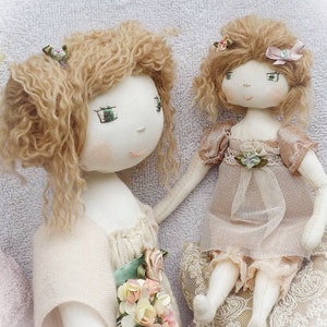 Pode incluir: Duas bonecas de tecido feitas à mão com cabelo castanho encaracolado. A boneca à esquerda está vestindo um vestido branco com uma faixa verde e um buquê de flores rosa e amarelas. A boneca à direita está vestindo um vestido bege com renda e um laço rosa no cabelo. As bonecas estão sentadas em uma superfície branca e fofa. O texto "www.VerityHope.com" é visível no fundo.